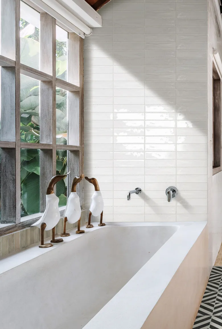 Ett ljust badrum där ett fristående badkar är placerat vid ett stort fönster som släpper in naturligt ljus. Väggarna är klädda med vita, glaserade kakelplattor i smalt format som ger en lugn och stilren känsla i rummet. På badkarskanten står dekorativa träfigurer och fönstrets trädetaljer bidrar till en varm och naturlig atmosfär.